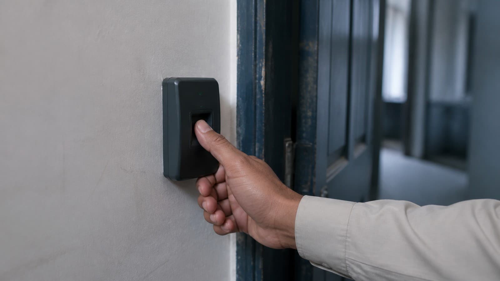 A hand pressing a finger onto a wall-mounted fingerprint access reader beside an office doorway