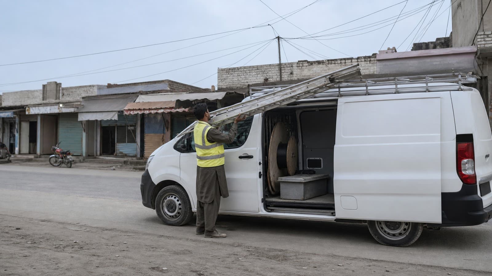 Technician lifting an aluminium ladder onto the roof rack of a service van, a drum of cable visible through the open side door