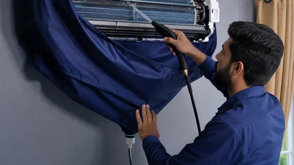 Technician washing down the indoor coil of a wall-mounted split AC, a service bag slung underneath to catch the runoff